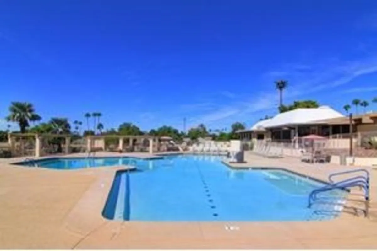 This exterior image showcases a sparkling swimming pool surrounded by lounge chairs and palm trees, with a nearby building suggesting a community clubhouse or amenity center. The setting is cheerful and inviting, with plenty of space for residents to swim, relax, or socialize under the sun.