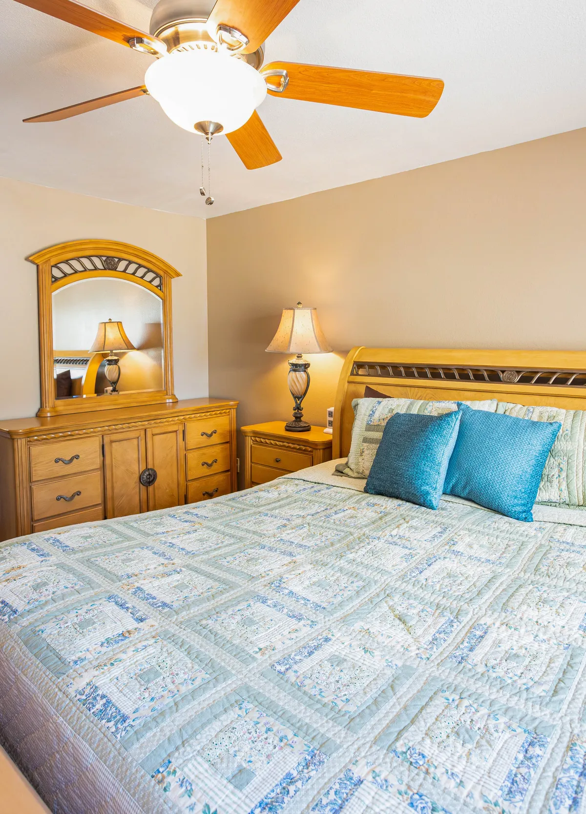 A welcoming bedroom scene with a quilted bedspread, decorative pillows, wooden furniture, and a lamp beside the bed. The natural lighting and tidy arrangement evoke a restful and inviting atmosphere, perfect for accommodating guests or family.