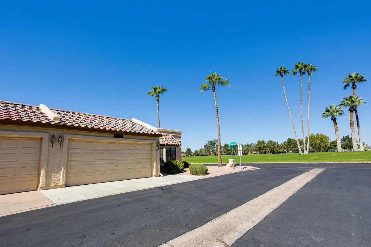 This image shows the exterior of the property, including palm trees, the driveway, and garage doors, all bathed in sunshine. The setting feels tranquil and mature, suggesting a relaxed, walkable neighborhood environment.