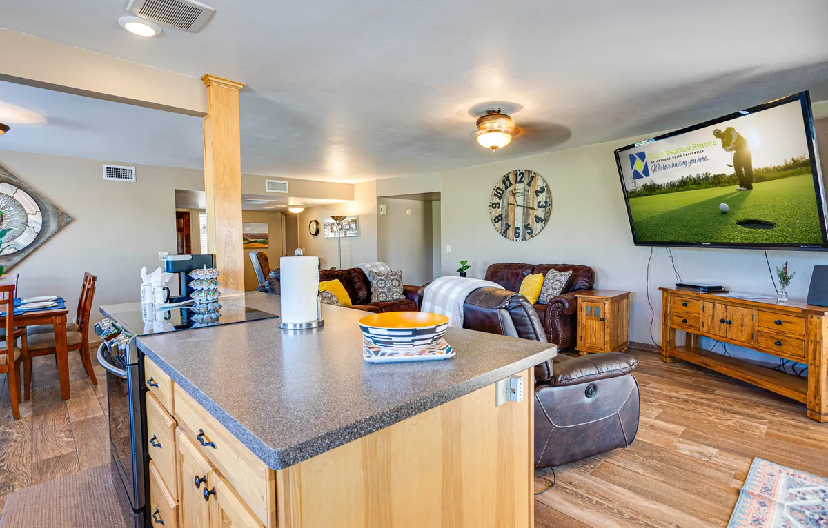 This image captures an inviting open-concept living area with a kitchen island, a large television prominently displayed, and multiple seating and dining options. The area is bathed in natural light, and the furnishings—including plush recliners—combine comfort with style. There are cues of relaxed social gatherings, from watching TV to sharing meals or enjoying casual chats.
