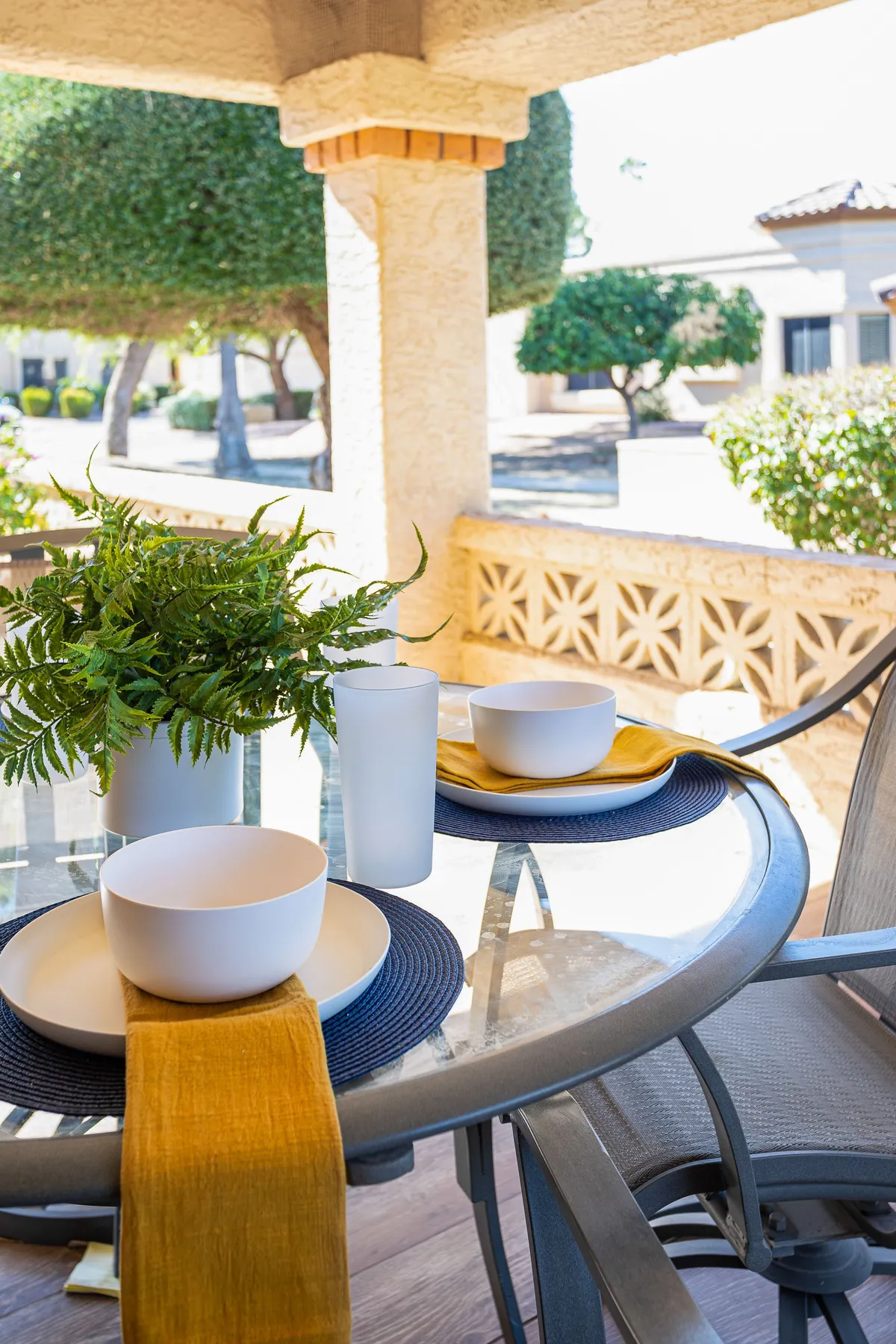 An inviting outdoor patio featuring a glass table set for dining with white bowls, a cheerful yellow table runner, and a green plant centerpiece. The setting suggests a tranquil and private space, ideal for relaxing or enjoying a meal outdoors with a pet nearby.
