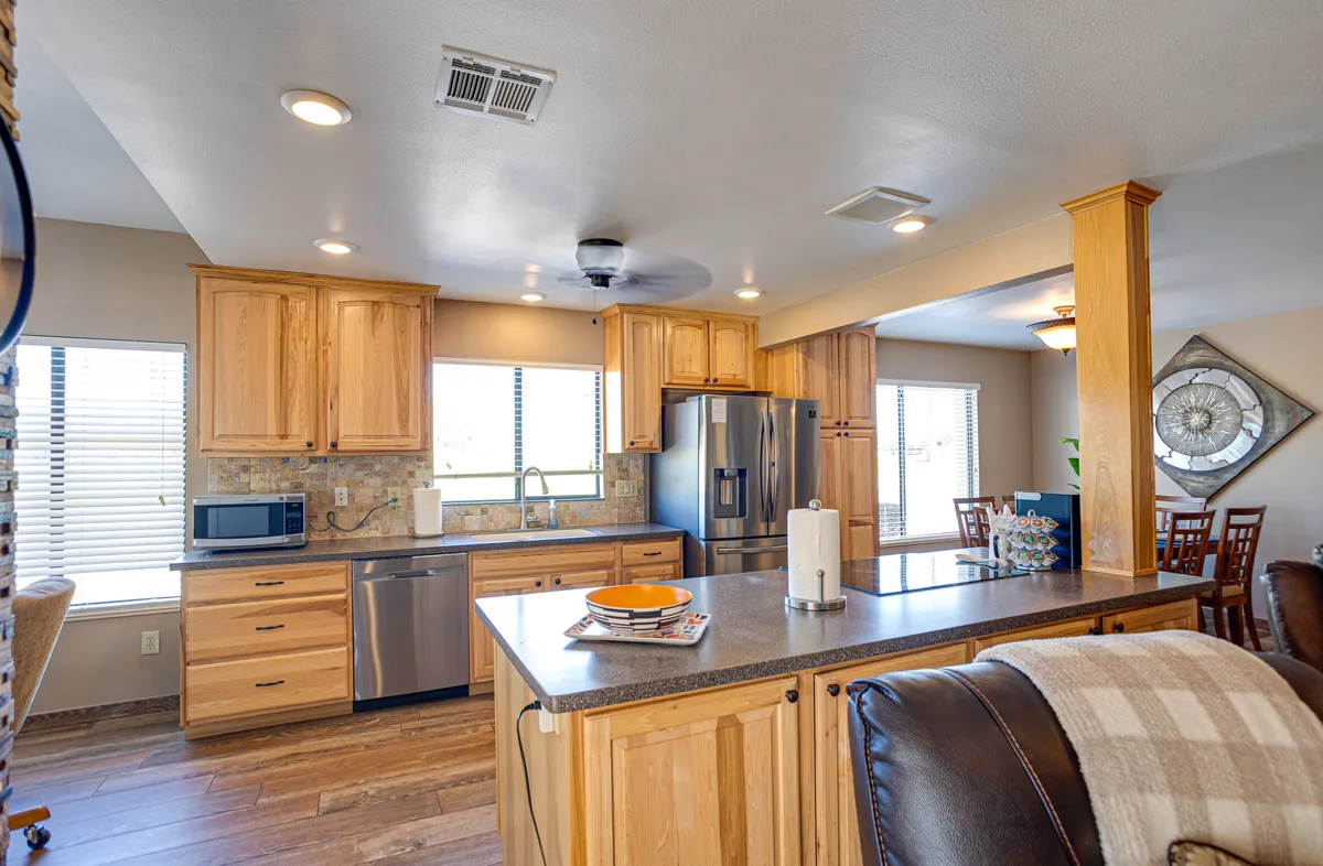 A wide view of an open kitchen with a central island, warm wooden cabinets, and stainless steel appliances, seamlessly flowing into a dining area with large windows. The layout invites both cooking and socializing, and the atmosphere is warm and welcoming.